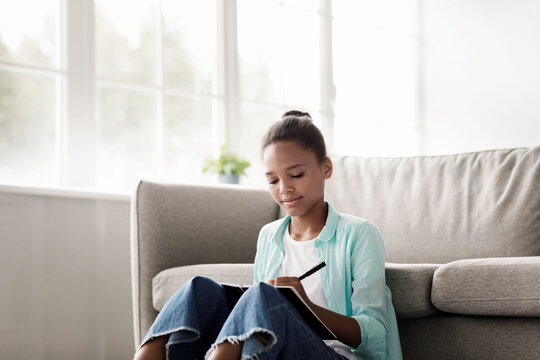 A concentrated teen girl sits on the floor in her room, taking notes in her notebook. She is focused on her studies, preparing for upcoming tests in a bright and inviting space. - Powered by Adobe