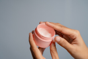 Close Up Of Hands Holding A Pink Open Jar Of Cream With A Light Pink Hue