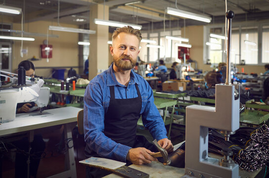 Work with leather products.Shoemaker. Portrait of a friendly man sitting at his workplace and using a hammer to make belts in a factory. Andult bearded craftsman sits in an apron and prepares - Powered by Adobe