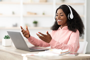 A young woman sits at a desk, wearing headphones, happily participating in an online class. She interacts with her laptop in a bright and cozy environment filled with plants.
