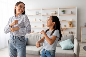 Fototapeta premium An excited young mom and her cute daughter dance together in their cozy living room, smiling and enjoying their favorite song. They share a fun and carefree moment, filled with laughter.