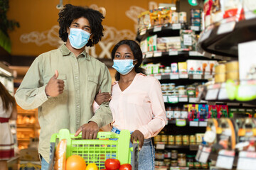 A young couple stands close together in a grocery store aisle, smiling while holding a shopping cart filled with fruits and vegetables. They are wearing face masks.