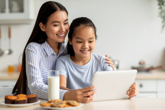 In a bright kitchen, a joyful Asian mother and her daughter are using a digital tablet to discover new recipes. They enjoy their time together, surrounded by baked goods and milk.