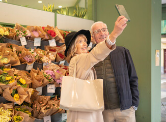 Happy senior couple smiling, using smartphone to take selfie in front of flower bouquets