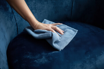 A close-up shot of a hand cleaning a blue velvet armchair with a microfiber cloth. The focus is on the gentle cleaning action on the luxurious upholstery. Furniture care. Home maintenance.