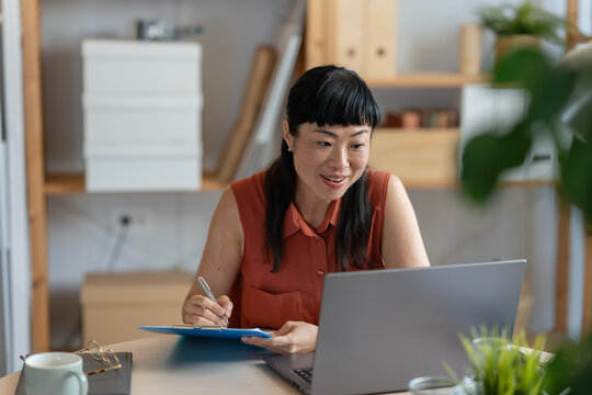 Smiling Asian woman entrepreneur writing notes while holding a coffee mug, working at her laptop in a calm and organized home office - Powered by Adobe