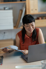 Asian woman small entrepreneur reviewing paperwork at her home office desk, working with focus...