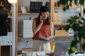 Asian woman small entrepreneur standing in her home office, smiling while talking on the phone and...
