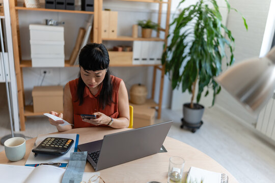 Asian woman entrepreneur in her home office, checking receipts and entering numbers on her phone while working with a laptop and calculator at a round desk