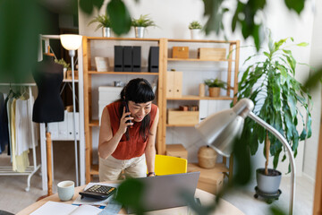 Asian woman small entrepreneur on a phone call in her bright home office, leaning over her desk...