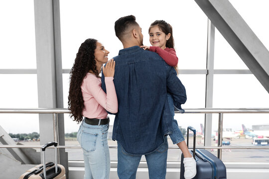 A family stands inside the airport, looking excited for their upcoming travel. The father holds his daughter on his shoulder while the mother smiles at them. Luggage is nearby.