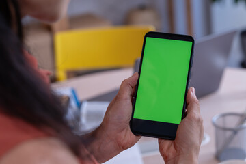 Asian woman small entrepreneur using a smartphone with a green screen mockup in her home office, checking digital content while managing her business tasks
