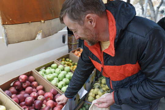 Man choosing fresh red apples at an outdoor farmers' market. Healthy grocery shopping with a reusable bag