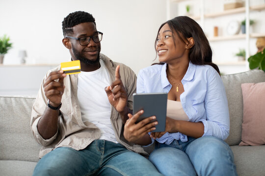 Young black couple sits on a cozy sofa discussing an online purchase. The woman looks excited while holding a tablet, and the man holds a credit card, signaling her to wait before making a buy.