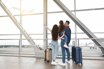 A family stands together in an airport, with luggage beside them. They look out a large window,...