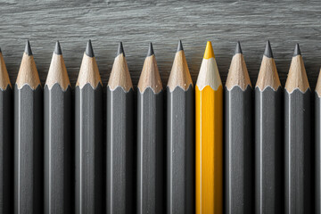 A sharp yellow pencil stands vertically in a row of identical dark gray pencils on a wood-grain surface. Strong symmetry. Individuality, uniqueness, leadership concept.