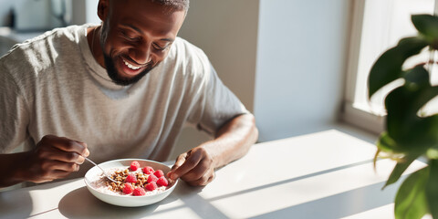A young Black man with a beard sits at a sunny table, smiling brightly while eating a bowl of oatmeal with raspberries. Healthy breakfast, joyful lifestyle, nutrition concept.