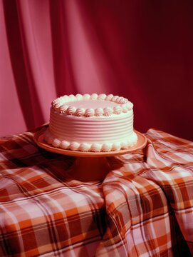 White cake on a red cake stand on a plaid tablecloth. the cake is round and has a ruffled design on top. the background is a solid red color, and the tablecloth is a warm orange and red plaid pattern.