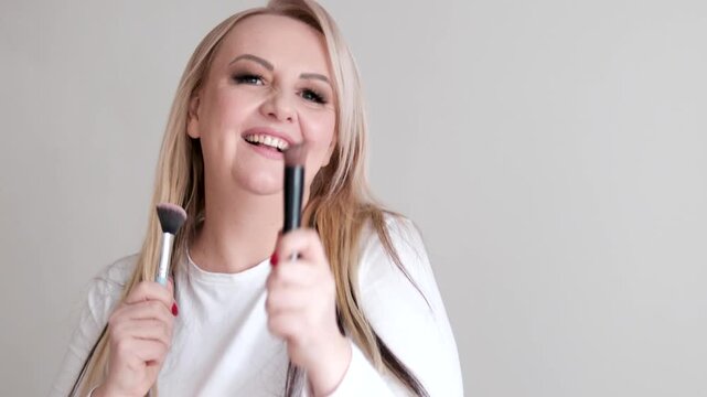 joyful European woman about forty five years old dances while holding makeup brushes in both hands smiling with red nails white top long lashes and long hair expressing happiness and lively beauty.