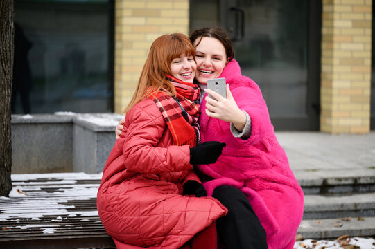 Two women take selfie while sitting on bench, wearing winter coats. Friends snap selfie on smartphone: warm hugs and bright smiles, tender, playful memories, sincere, joyful atmosphere.