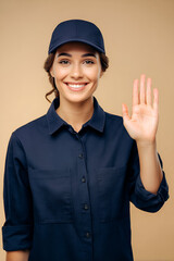 Smiling young woman waves hello wearing a dark uniform and cap against a neutral background