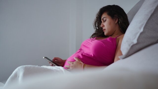 Pregnant woman relaxing in bed, using smartphone and touching her baby bump
