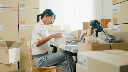 Youth attractive Asia female entrepreneur small business check parcel box key barcode number on cardboard box in laptop computer check inventory product ready for shipment at small online retail shop. © tirachard