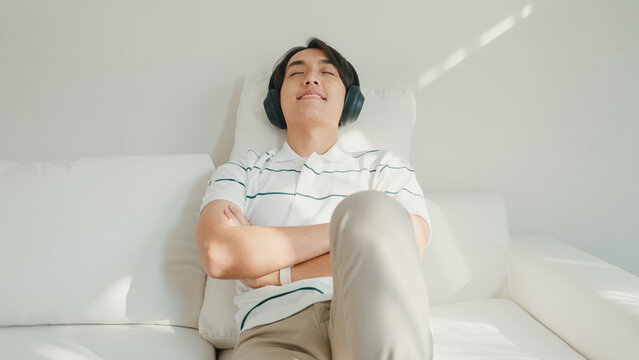 Youth attractive Asia male with casual cloth calm feeling lay down on sofa close eyes wear headphone listen music playlist relaxing at living room in cozy house. Lifestyle leisure at home concept.