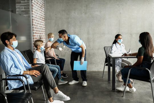 Middle eastern young man doctor greeting teen boy patient with elbow bump, multiracial group of people sitting at clinic, waiting for vaccination against COVID-19, wearing protective face masks - Powered by Adobe