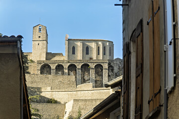 Traditional village scene in Sisteron, France, featuring beige stucco buildings and tiled roofs. A historic stone church with arched windows and a bell tower rises above the town
