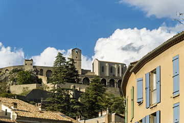 Traditional village scene in Sisteron, France, featuring beige stucco buildings and tiled roofs. A historic stone church with arched windows and a bell tower rises above the town
