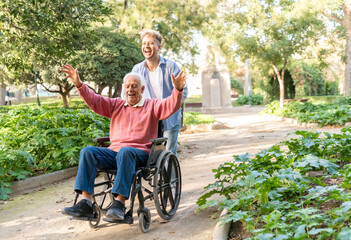 Young adult man laughing while pushing cheerful grandfather senior man in wheelchair in park