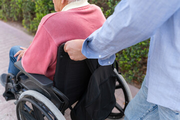 Close-up of male unrecognizable caregiver pushing elderly man in wheelchair outdoors
