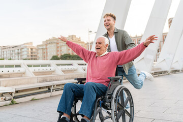 Laughing grandson pushing cheerful elderly man in wheelchair with arms raised on urban bridge