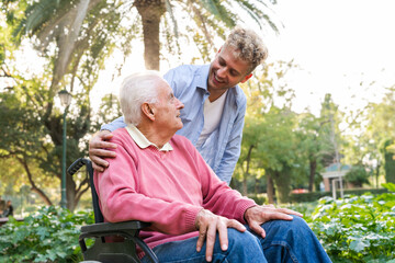 Grandson standing and smiling at senior man in wheelchair with hand on his shoulder at park