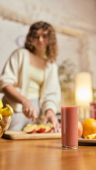 Fresh fruit smoothie glass in foreground with woman preparing ingredients in cozy kitchen. Concept of healthy recipe tutorials, nutrition blogs, wellness meal prep, smoothie brand promos.