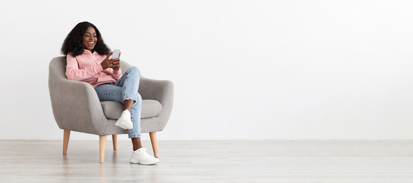 A woman sits comfortably in a gray chair, wearing casual clothes and white sneakers. She smiles while looking at her smartphone in a simple, well-lit room.