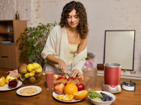 Woman cutting fruit for smoothie surrounded by colorful produce and kitchen tools. Concept of food-prep visuals, nutrition workshops, smoothie brand ads, healthy living materials.