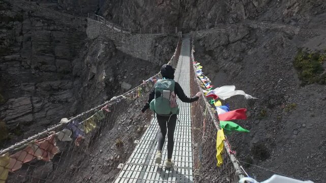 Wide Shot of Woman Hiker Crossing Mountain Suspension Bridge with Prayer Flags at Annapurna circuit trek on the way to Tilicho lake.