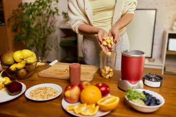 Woman adding chopped fruit into blender cup while preparing homemade smoothie. Concept of healthy meal prep guides, recipe tutorials, nutrition storytelling, wellness branding.