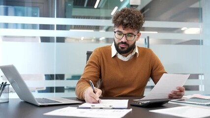 Busy businessman fills out documents with a pensitting at a work desk at workplace in modern glass business office. Financier in casual sweater doing paperwork, financial report, engaged in accounting - Powered by Adobe