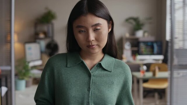 Tilt up portrait of confident Asian businesswoman holding document folder looking at camera while working in modern office