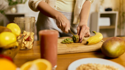 Hands slicing ripe kiwi for smoothie preparation with fruits and blender on table. Concept of recipe steps, short-form cooking videos, nutrition blogs, healthy lifestyle materials.