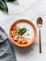 Bowl of granola with a spoon on a white marble countertop. the granola is made up of oats, nuts, and dried fruit, and there is a small green leaf on top of the granola.