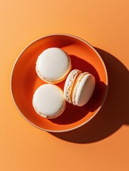 Three white macarons in an orange bowl. the bowl is placed on an orange background, casting a shadow on the right side of the image.