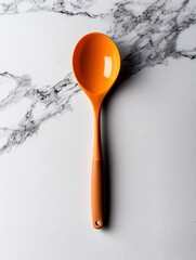 Orange ladle lying on a white marble surface. the ladle has a long handle with a small hole in the center. the handle is made of a shiny orange material and appears to be made of wood.