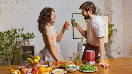 Couple cheering with green and pink smoothies in bright kitchen environment. Concept of healthy lifestyle campaigns, beverage product promotion, social media wellness content, nutrition marketing.