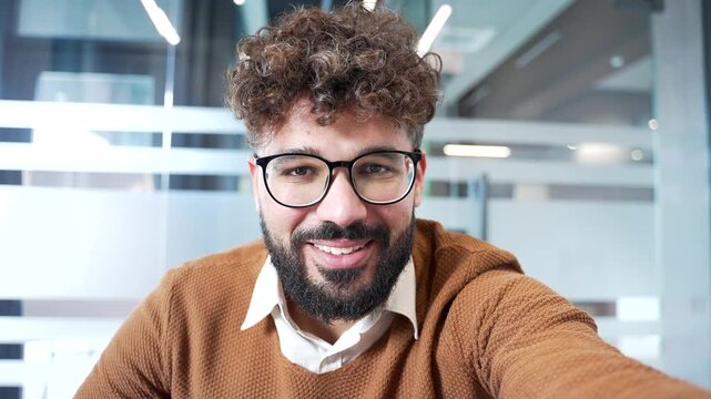 POV Smiling businessman talking on video call using a phone in the office. Confident male professional greeting waving hand looking at camera. Online meeting with client or business partner. Close up
