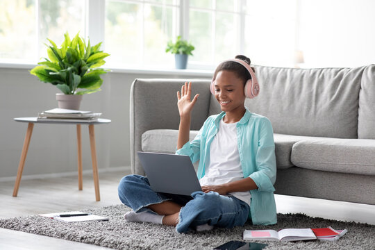 A teenager girl is sitting on the floor, wearing headphones and greeting her teacher during a video call. She is studying from home, surrounded by books and a cozy setting.