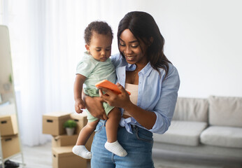 A young African American woman holds her baby while smiling at her cellphone. They are enjoying educational content together in a cozy living room filled with boxes.